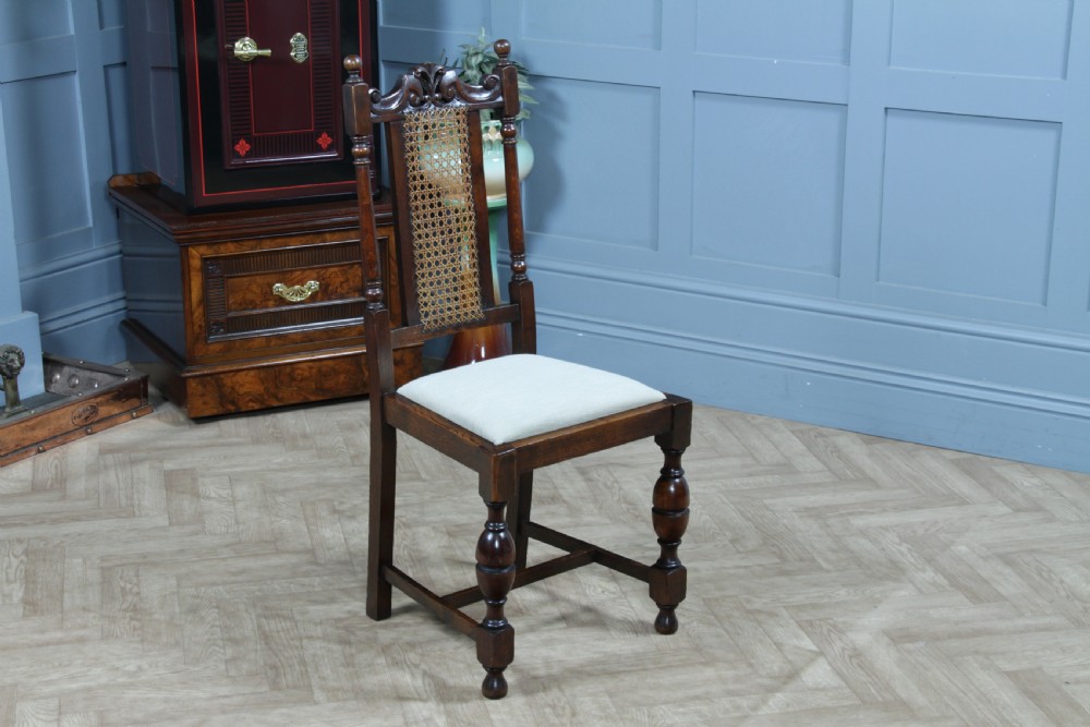 A set of four dark oak Jacobean Revival dining chairs featuring woven cane backs, carved crests, turned legs, and cream fabric seats. View XIX