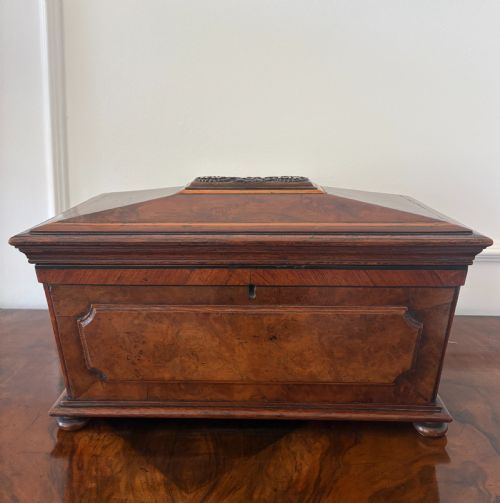 fig 2. A Victorian burr walnut sarcophagus-shaped tea caddy featuring a carved top plaque, stepped lid, keyhole escutcheon, and small bun feet.