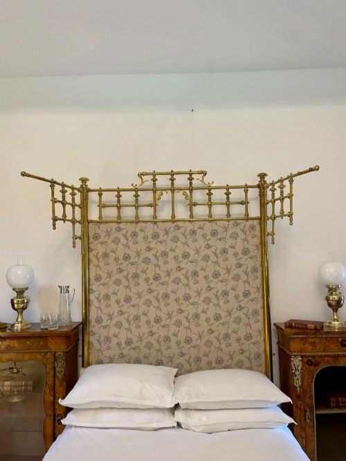 fig 3. An antique Victorian solid brass half tester double bed featuring turned posts, spindle balusters, cast decorative mounts, and an upholstered floral panel.