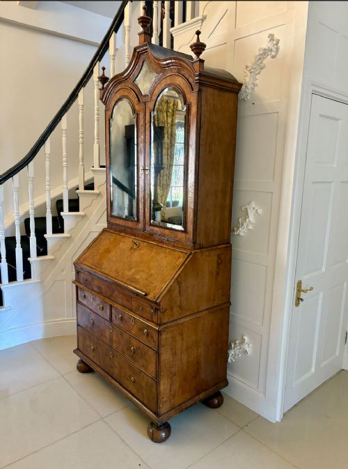 An antique Queen Anne burr walnut bureau bookcase featuring a double-domed cresting with urn finials, double mirrored doors, a sloping fall-front desk section, and a chest of drawers on bun feet. View VI