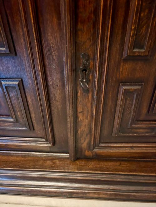 fig 2. A dark carved oak sideboard featuring geometric panelled cupboard doors with central lion mask carvings and a detailed relief frieze.