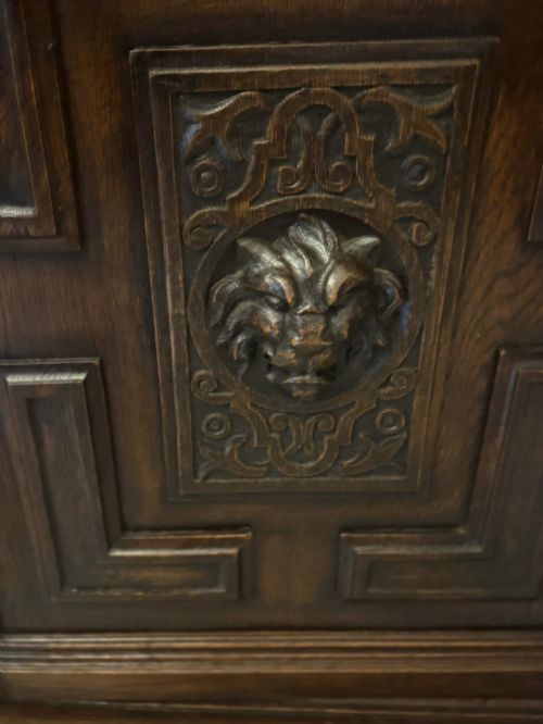 fig 3. A dark carved oak sideboard featuring geometric panelled cupboard doors with central lion mask carvings and a detailed relief frieze.