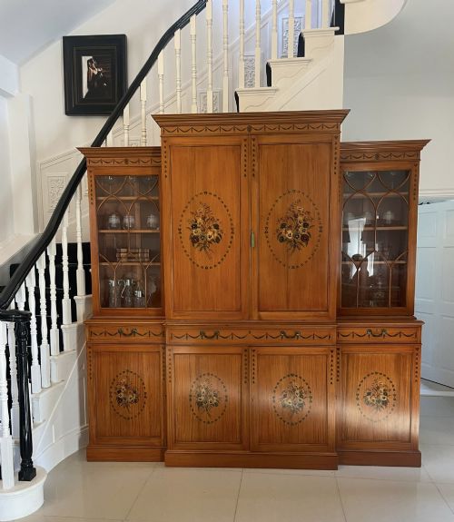 fig 1. A large satinwood breakfront bookcase featuring hand-painted floral cartouches, upper glazed display doors, and lower cupboard panels.
