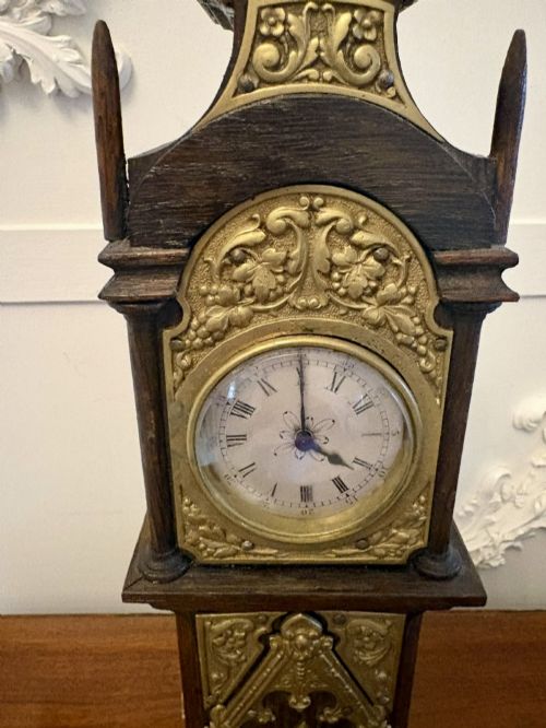 fig 3. A dark oak miniature grandfather table clock with ornate embossed brass panels and a white dial featuring Roman numerals.