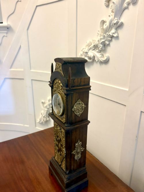 fig 5. A dark oak miniature grandfather table clock with ornate embossed brass panels and a white dial featuring Roman numerals.
