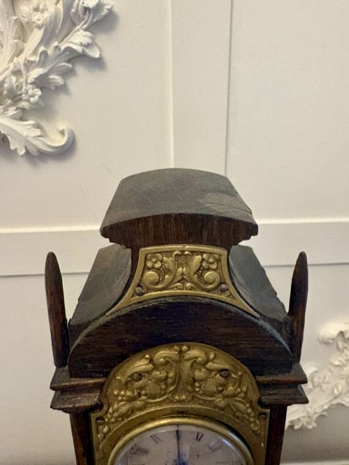 fig 7. A dark oak miniature grandfather table clock with ornate embossed brass panels and a white dial featuring Roman numerals.