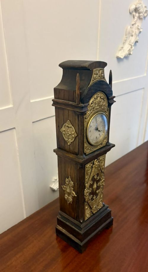 fig 8. A dark oak miniature grandfather table clock with ornate embossed brass panels and a white dial featuring Roman numerals.