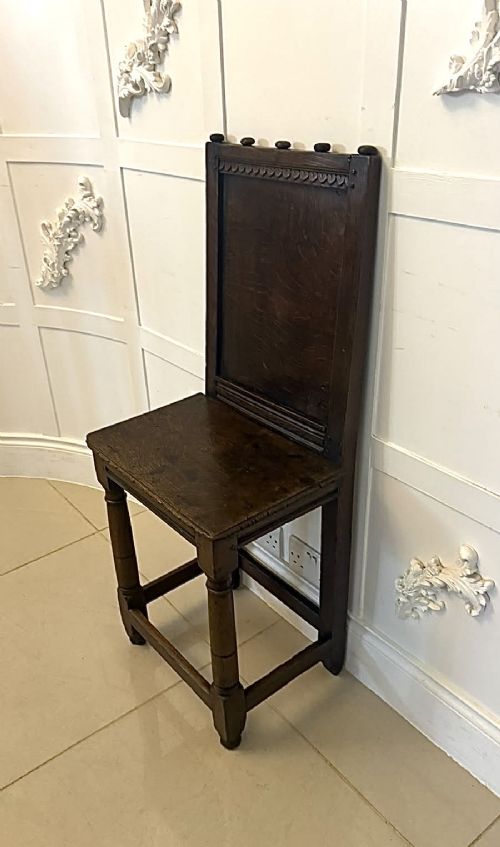 A dark oak 17th-century correction chair featuring a tall panelled backrest, carved crest rail with small wooden finials, solid wooden seat, turned front legs, and plain stretchers. View XI