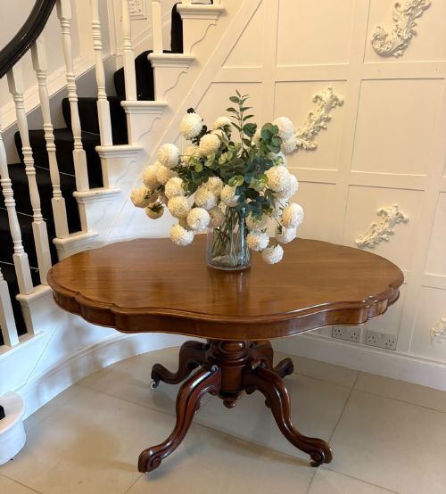 fig 2. A Victorian figured mahogany tilt-top centre table featuring a shaped serpentine top positioned vertically on a carved central pedestal with four splayed legs.