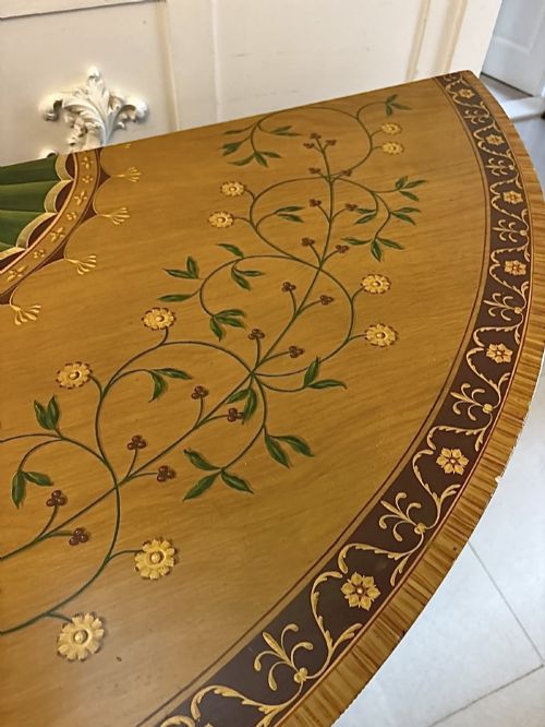 fig 5. A semicircular satinwood demi-lune cabinet featuring hand-painted neoclassical leaf swags, a central figural medallion, and short tapered feet.