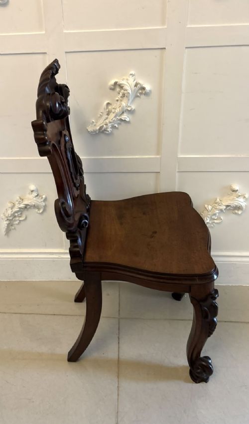 A pair of Victorian carved mahogany hall chairs with shaped timber seats, scrolled cartouche backrests, and carved cabriole legs. View VII