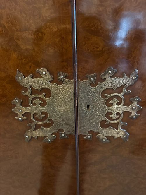 fig 1. An antique burr walnut cocktail cabinet featuring decorative brass hardware, carved scroll cresting, two cupboard sections, and cabriole legs.