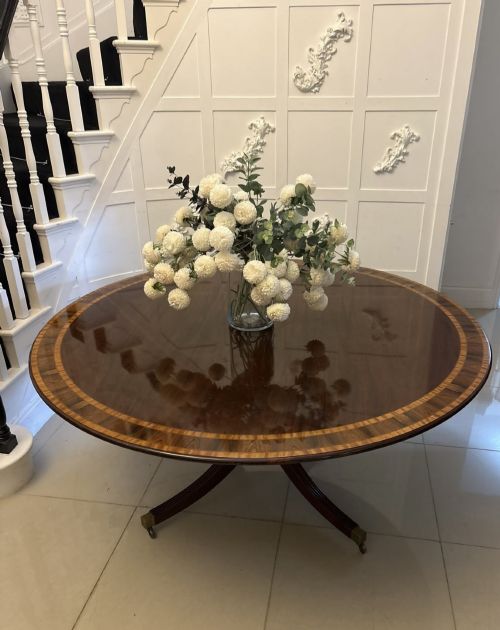 fig 3. A large circular mahogany dining table featuring rosewood and satinwood crossbanding set upon a central pedestal base with reeded legs ending in brass castors.