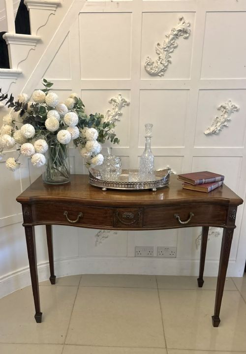 An Edwardian mahogany console table featuring a serpentine front, carved central motif, brass handles, and square tapered legs. View VI