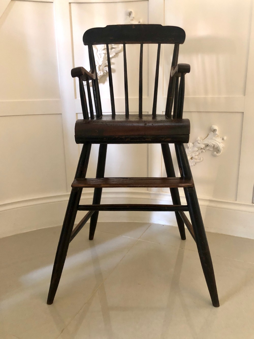 A dark-stained wooden antique child high chair featuring a vertical spindle backrest, armrests, splayed legs, and a built-in wooden footrest.