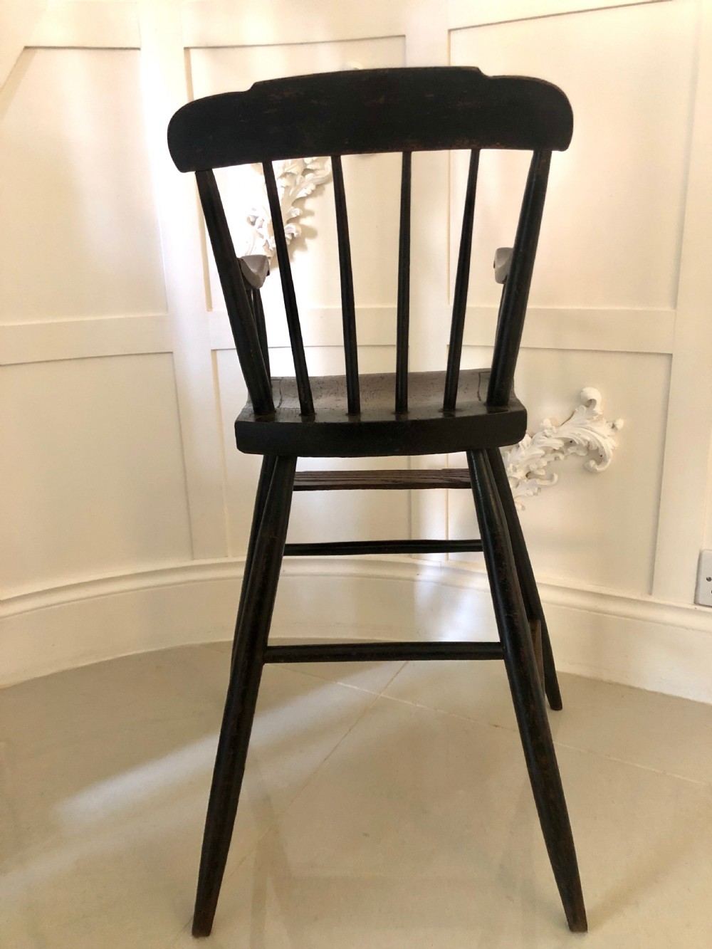 A dark-stained wooden antique child high chair featuring a vertical spindle backrest, armrests, splayed legs, and a built-in wooden footrest. View III