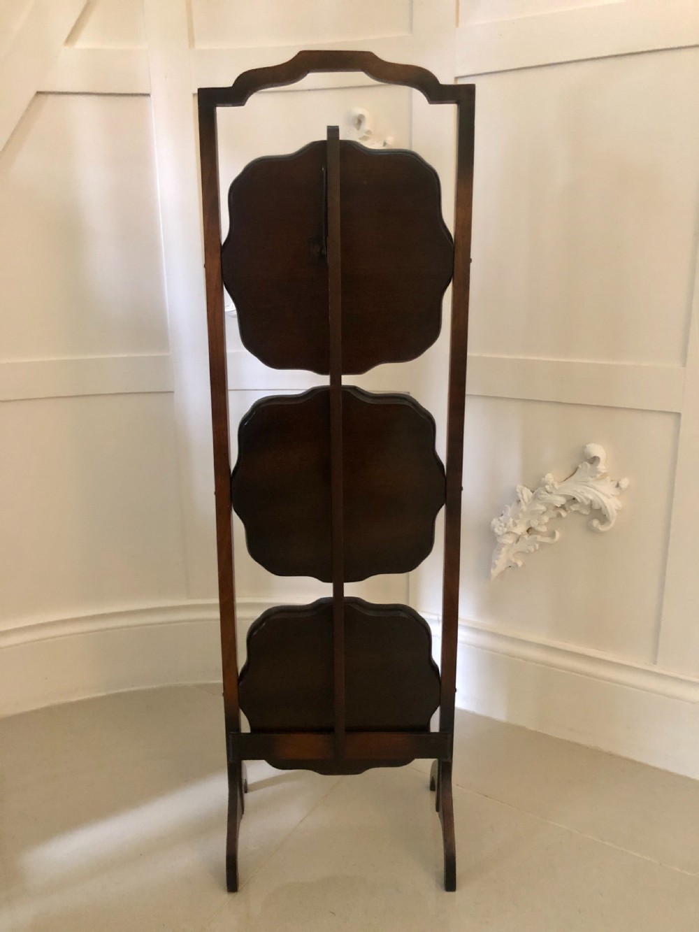 An antique three-tier walnut folding cake stand featuring shaped moulded trays, a curved top carrying handle, and straight wooden side supports. View II