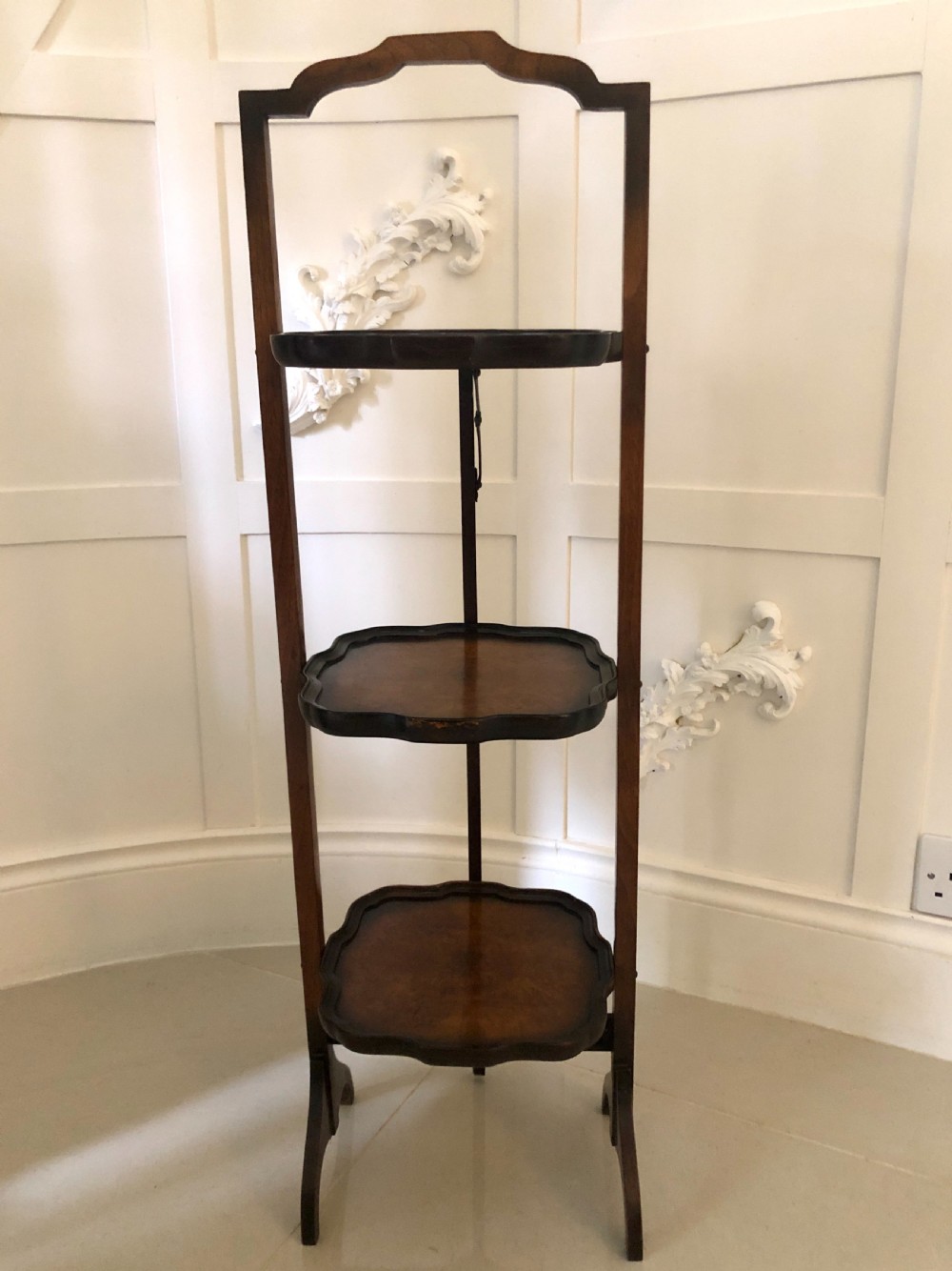 An antique three-tier walnut folding cake stand featuring shaped moulded trays, a curved top carrying handle, and straight wooden side supports. View III