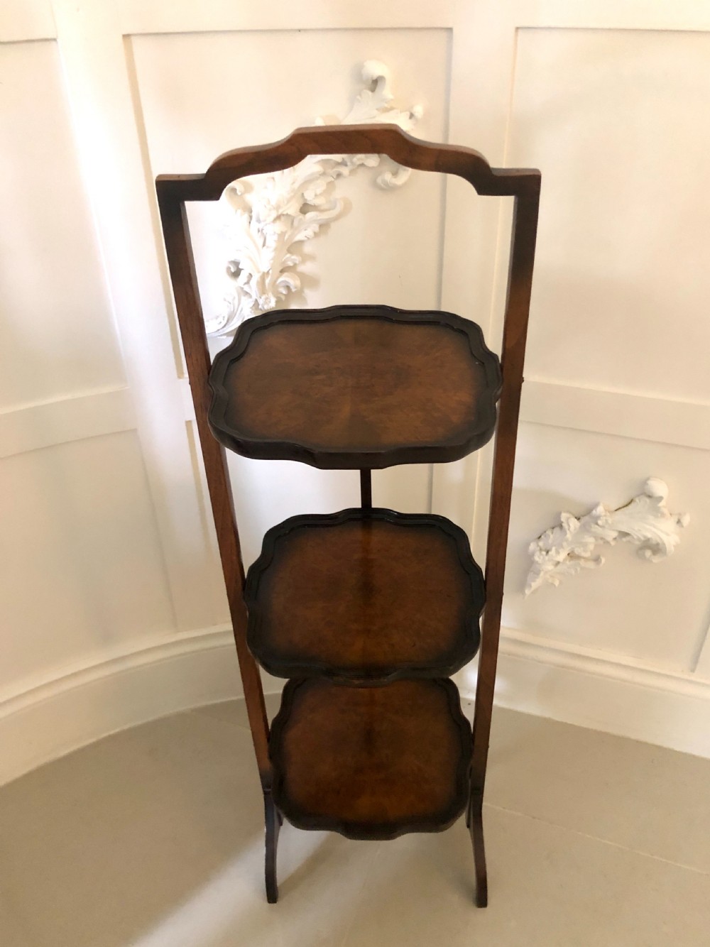 An antique three-tier walnut folding cake stand featuring shaped moulded trays, a curved top carrying handle, and straight wooden side supports. View V