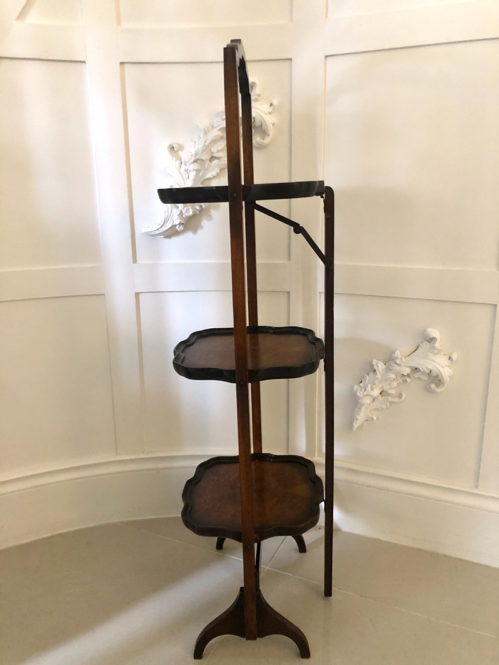 An antique three-tier walnut folding cake stand featuring shaped moulded trays, a curved top carrying handle, and straight wooden side supports. View IX
