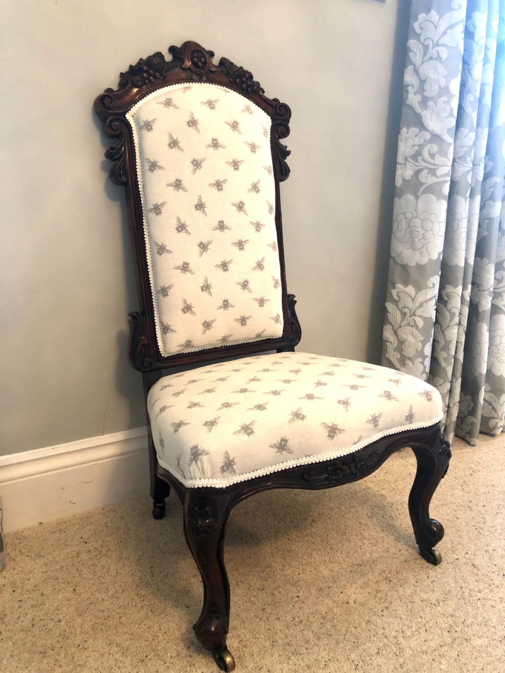 A Victorian carved rosewood ladies chair featuring a high upholstered backrest and seat with a grey bee pattern on cream fabric, raised on cabriole front legs with brass castors.