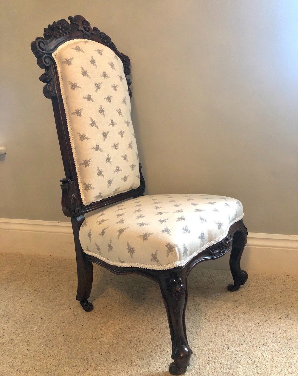 A Victorian carved rosewood ladies chair featuring a high upholstered backrest and seat with a grey bee pattern on cream fabric, raised on cabriole front legs with brass castors. View II
