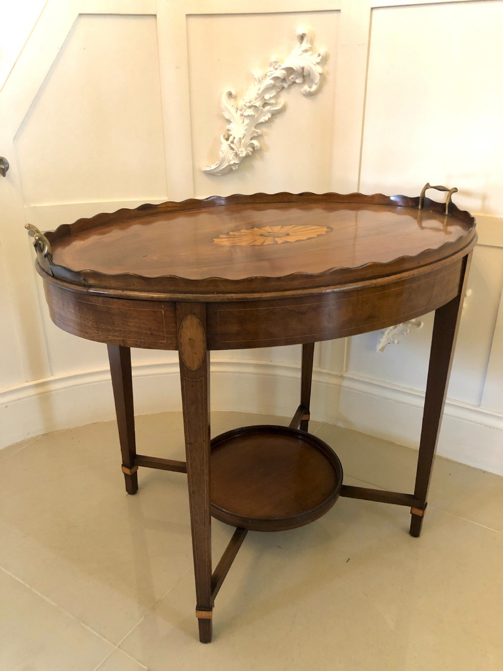 An Edwardian oval mahogany tray table featuring a scalloped gallery edge, brass side handles, satinwood inlay, and a lower circular shelf tier. View V