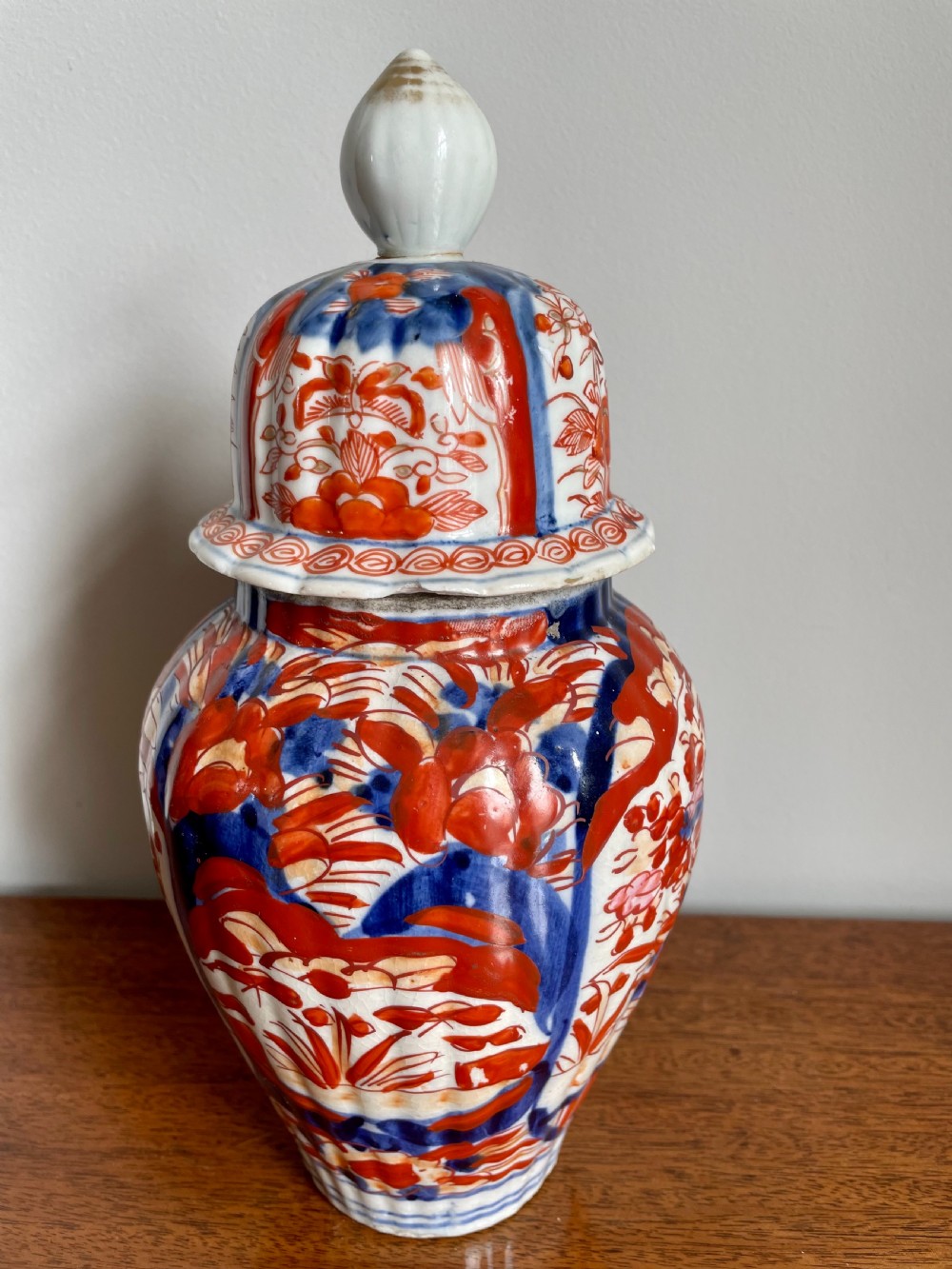 A fluted ceramic Imari vase with a matching lid, decorated in hand-painted floral patterns of iron-red and cobalt blue on a white background. View VII