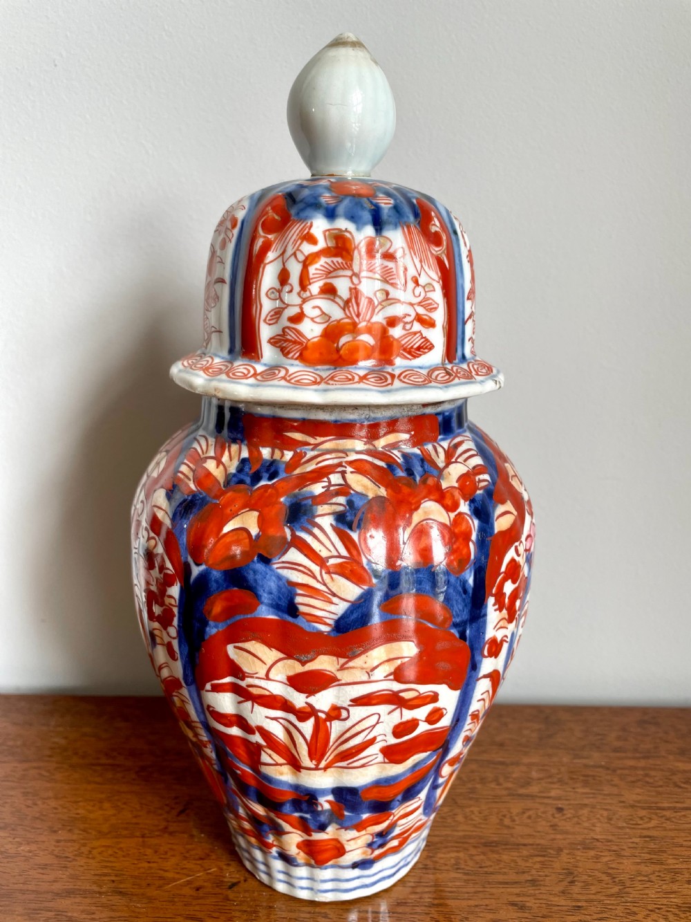 A fluted ceramic Imari vase with a matching lid, decorated in hand-painted floral patterns of iron-red and cobalt blue on a white background. View IX