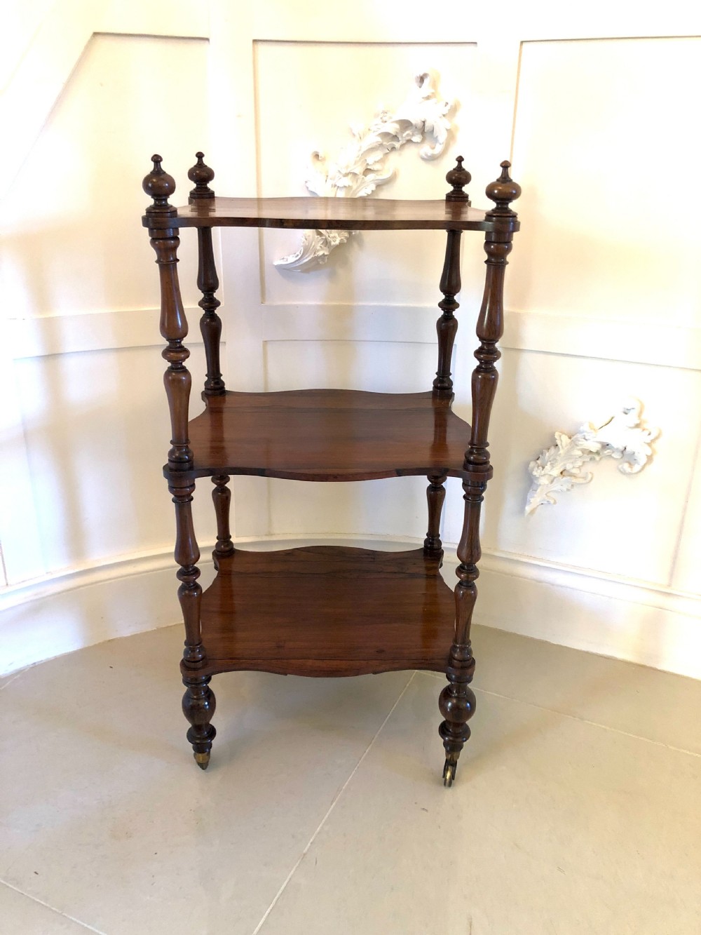 A three-tiered Victorian rosewood whatnot featuring serpentine shelf edges, turned corner supports, decorative finials, and brass castors.