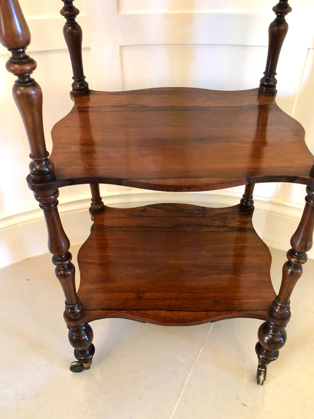 A three-tiered Victorian rosewood whatnot featuring serpentine shelf edges, turned corner supports, decorative finials, and brass castors. View V