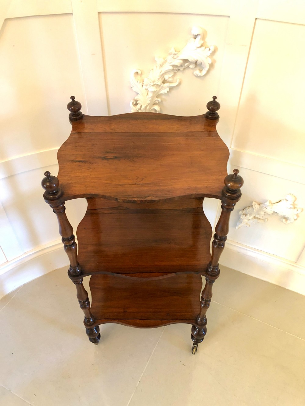 A three-tiered Victorian rosewood whatnot featuring serpentine shelf edges, turned corner supports, decorative finials, and brass castors. View IX