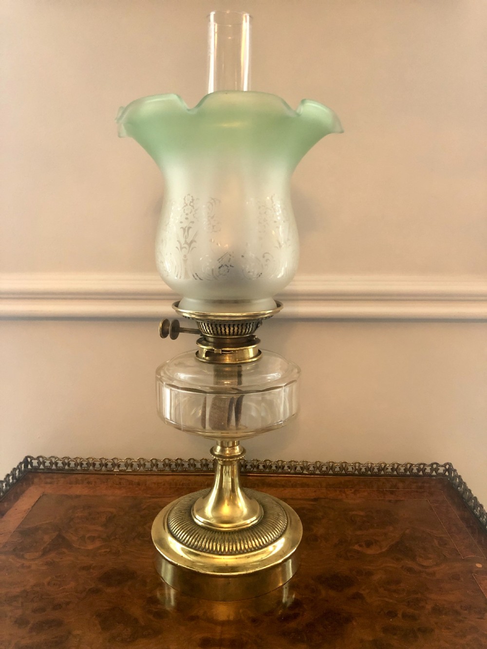 A Victorian oil lamp featuring a weighted brass base, clear glass reservoir, etched glass shade with a green crimped rim, and a tall clear glass chimney.