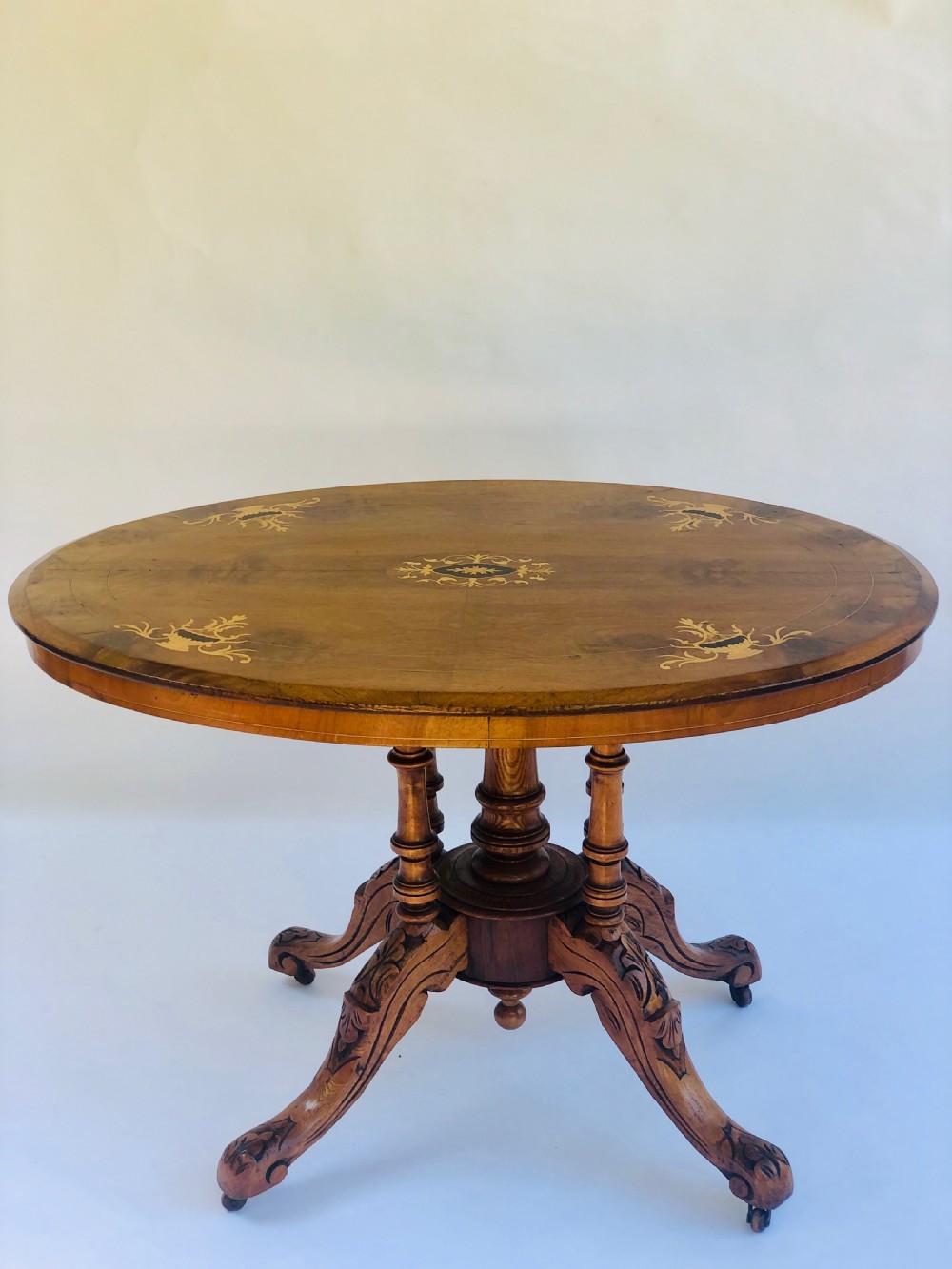 An oval Victorian walnut centre table featuring floral marquetry inlays on the top, supported by turned central columns and four carved legs on castors.