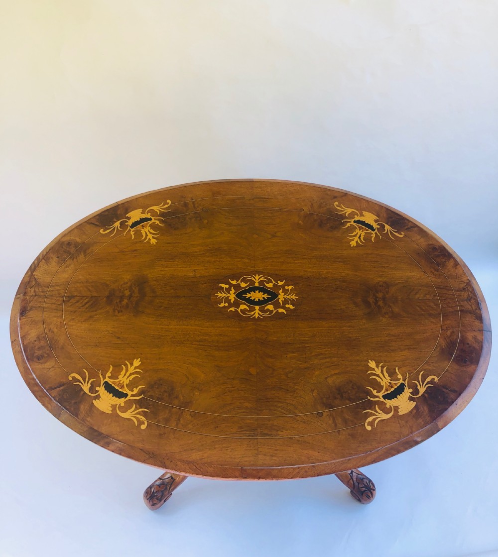 An oval Victorian walnut centre table featuring floral marquetry inlays on the top, supported by turned central columns and four carved legs on castors. View II