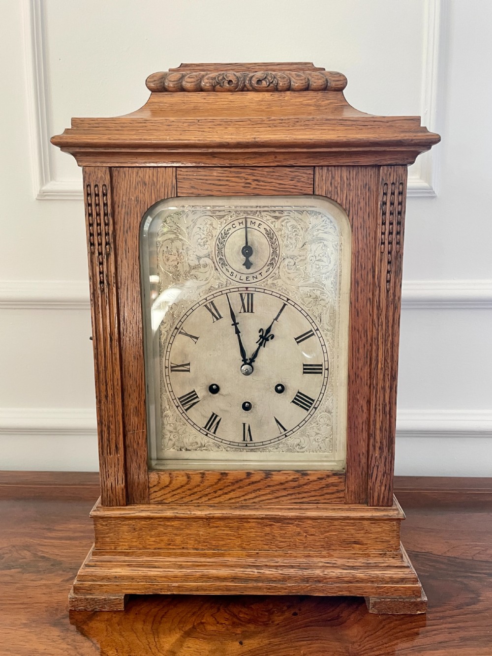 A Victorian carved oak bracket clock featuring a silvered engraved dial with Roman numerals, a chime-silent secondary dial, and carved gadrooned detailing along the top base.