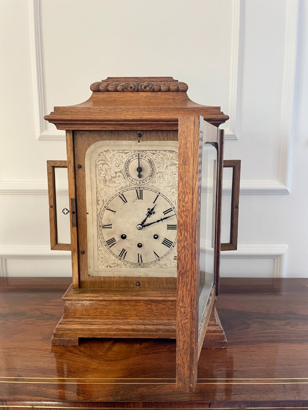 A Victorian carved oak bracket clock featuring a silvered engraved dial with Roman numerals, a chime-silent secondary dial, and carved gadrooned detailing along the top base. View II