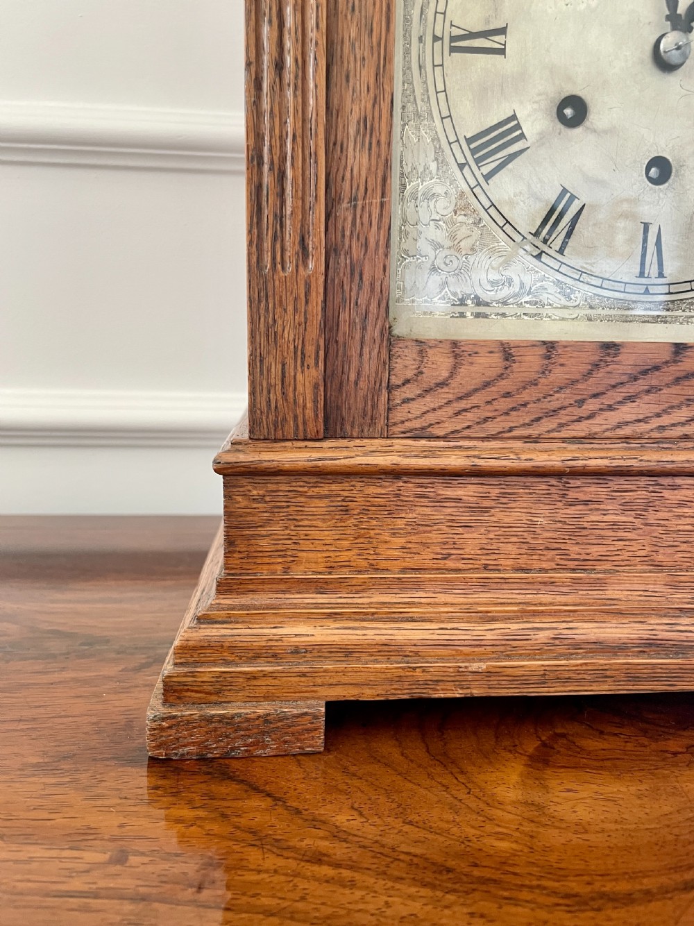 A Victorian carved oak bracket clock featuring a silvered engraved dial with Roman numerals, a chime-silent secondary dial, and carved gadrooned detailing along the top base. View XI
