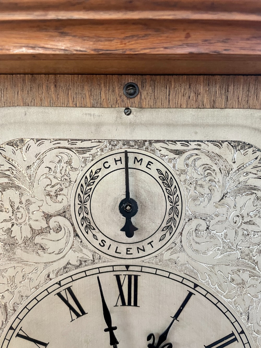 A Victorian carved oak bracket clock featuring a silvered engraved dial with Roman numerals, a chime-silent secondary dial, and carved gadrooned detailing along the top base. View XIV