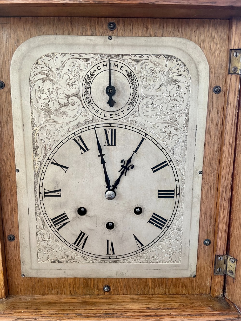 A Victorian carved oak bracket clock featuring a silvered engraved dial with Roman numerals, a chime-silent secondary dial, and carved gadrooned detailing along the top base. View XV