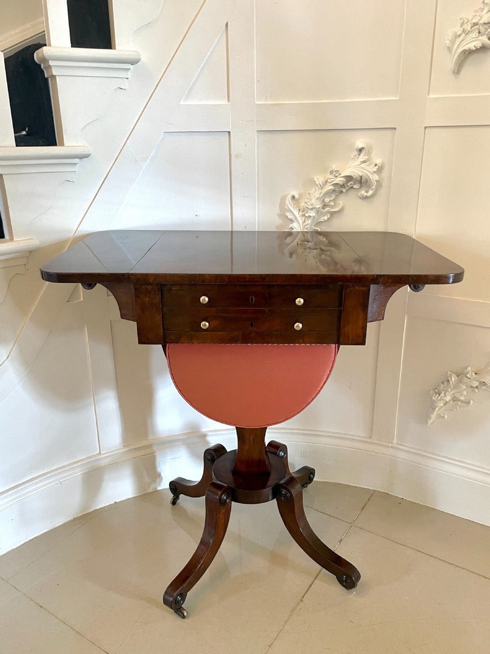 An antique Regency rosewood sewing table featuring small drop leaves, drawers with brass handles, a terracotta fabric work basket below, and a pedestal base with four splayed legs on castors. View V