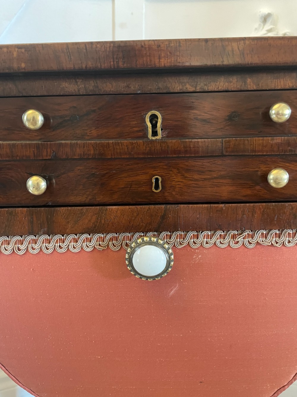 An antique Regency rosewood sewing table featuring small drop leaves, drawers with brass handles, a terracotta fabric work basket below, and a pedestal base with four splayed legs on castors. View IX