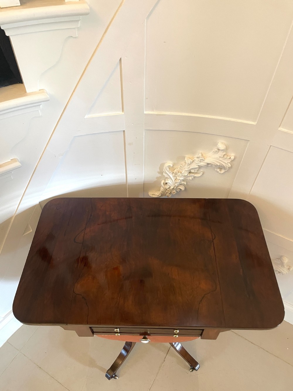 An antique Regency rosewood sewing table featuring small drop leaves, drawers with brass handles, a terracotta fabric work basket below, and a pedestal base with four splayed legs on castors. View X