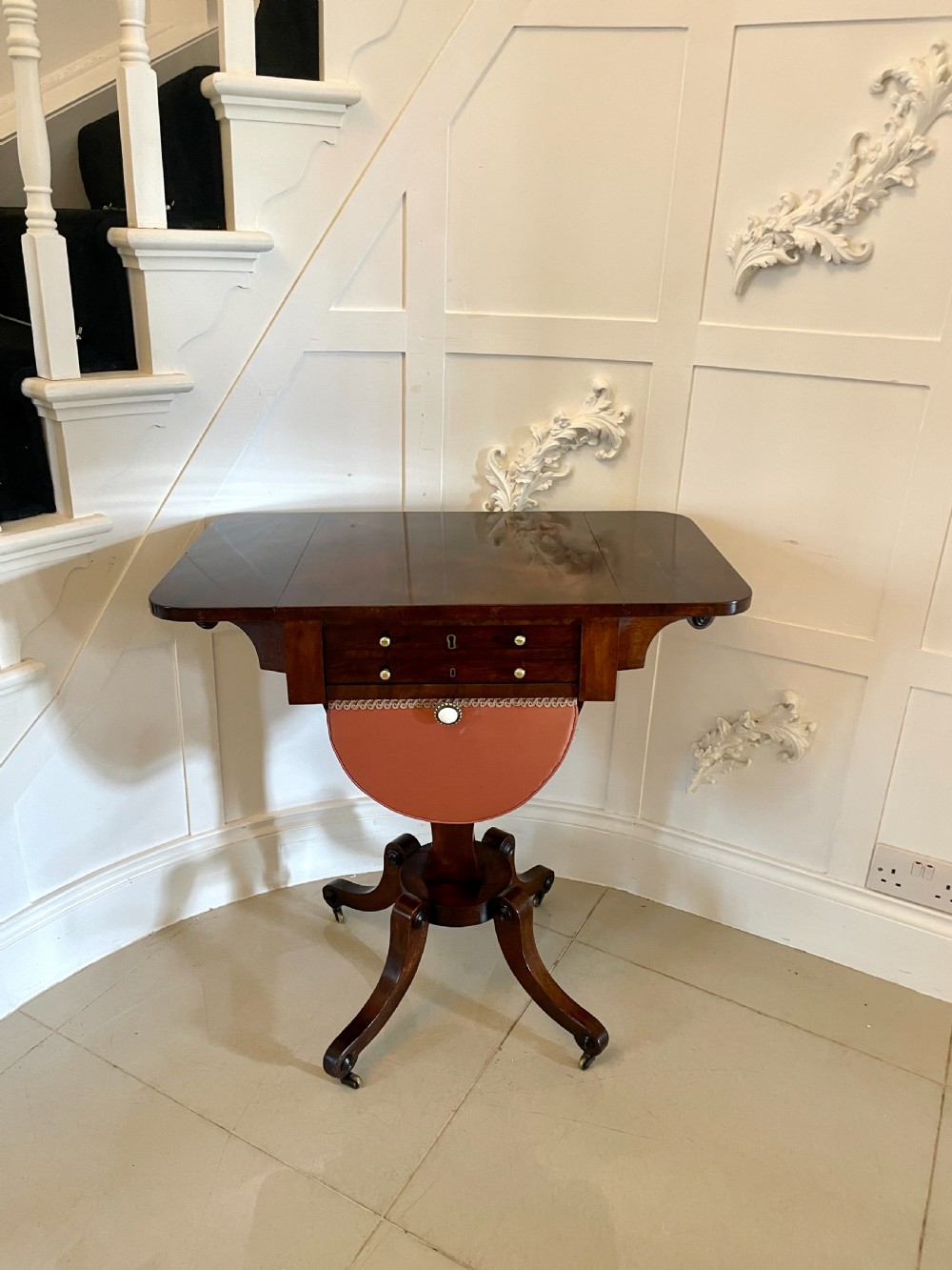 An antique Regency rosewood sewing table featuring small drop leaves, drawers with brass handles, a terracotta fabric work basket below, and a pedestal base with four splayed legs on castors. View XII