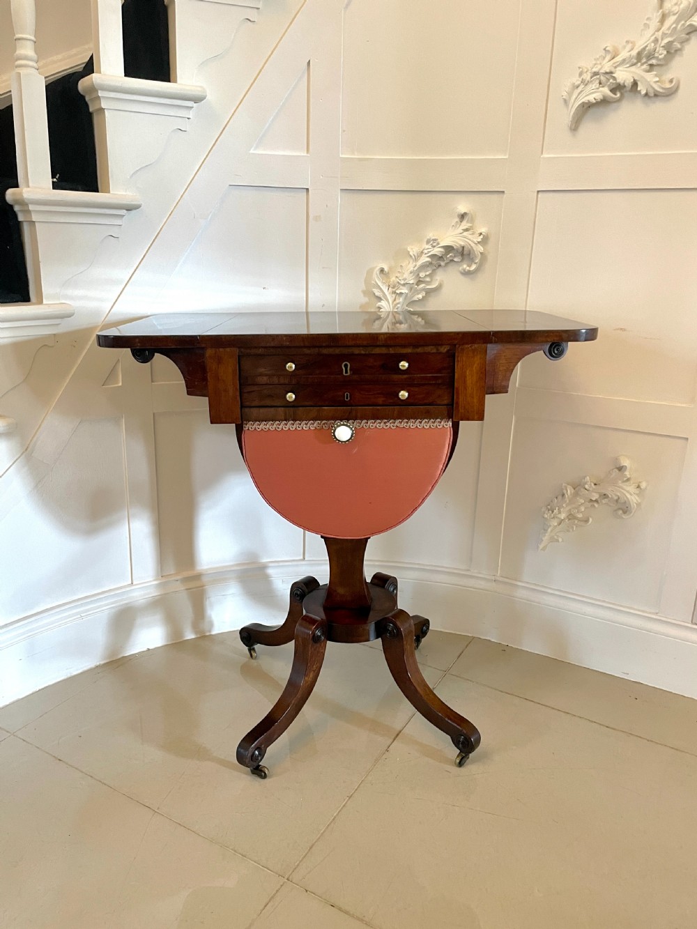 An antique Regency rosewood sewing table featuring small drop leaves, drawers with brass handles, a terracotta fabric work basket below, and a pedestal base with four splayed legs on castors. View XIII