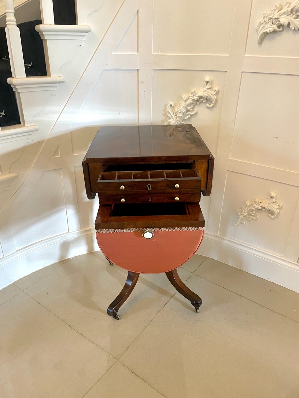 An antique Regency rosewood sewing table featuring small drop leaves, drawers with brass handles, a terracotta fabric work basket below, and a pedestal base with four splayed legs on castors. View XIV
