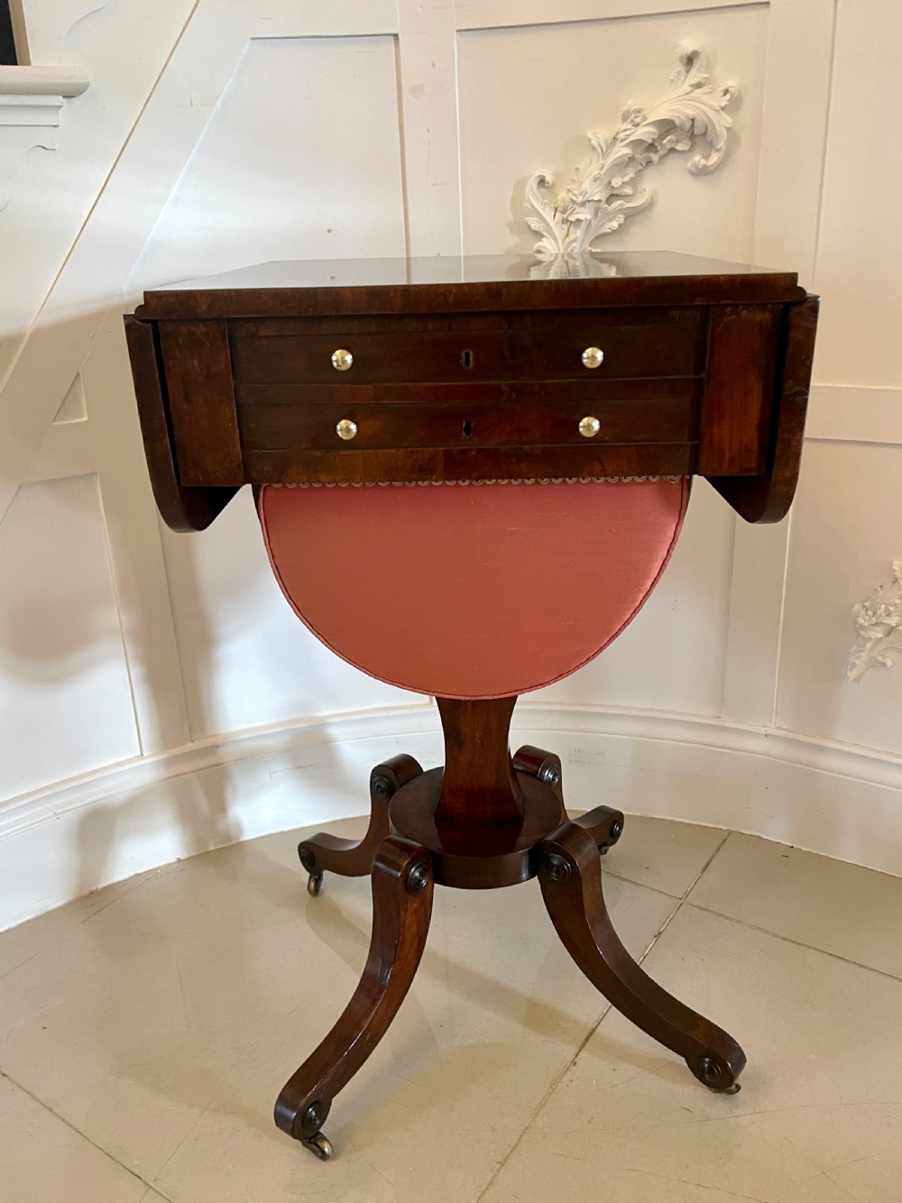 An antique Regency rosewood sewing table featuring small drop leaves, drawers with brass handles, a terracotta fabric work basket below, and a pedestal base with four splayed legs on castors. View XV