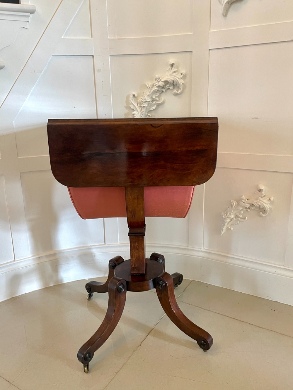 An antique Regency rosewood sewing table featuring small drop leaves, drawers with brass handles, a terracotta fabric work basket below, and a pedestal base with four splayed legs on castors. View XVI