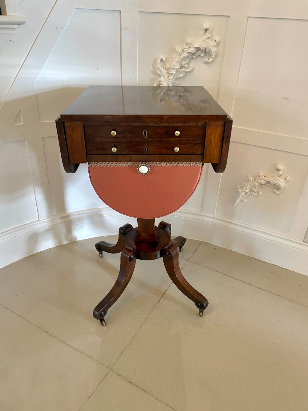 An antique Regency rosewood sewing table featuring small drop leaves, drawers with brass handles, a terracotta fabric work basket below, and a pedestal base with four splayed legs on castors. View XVIII