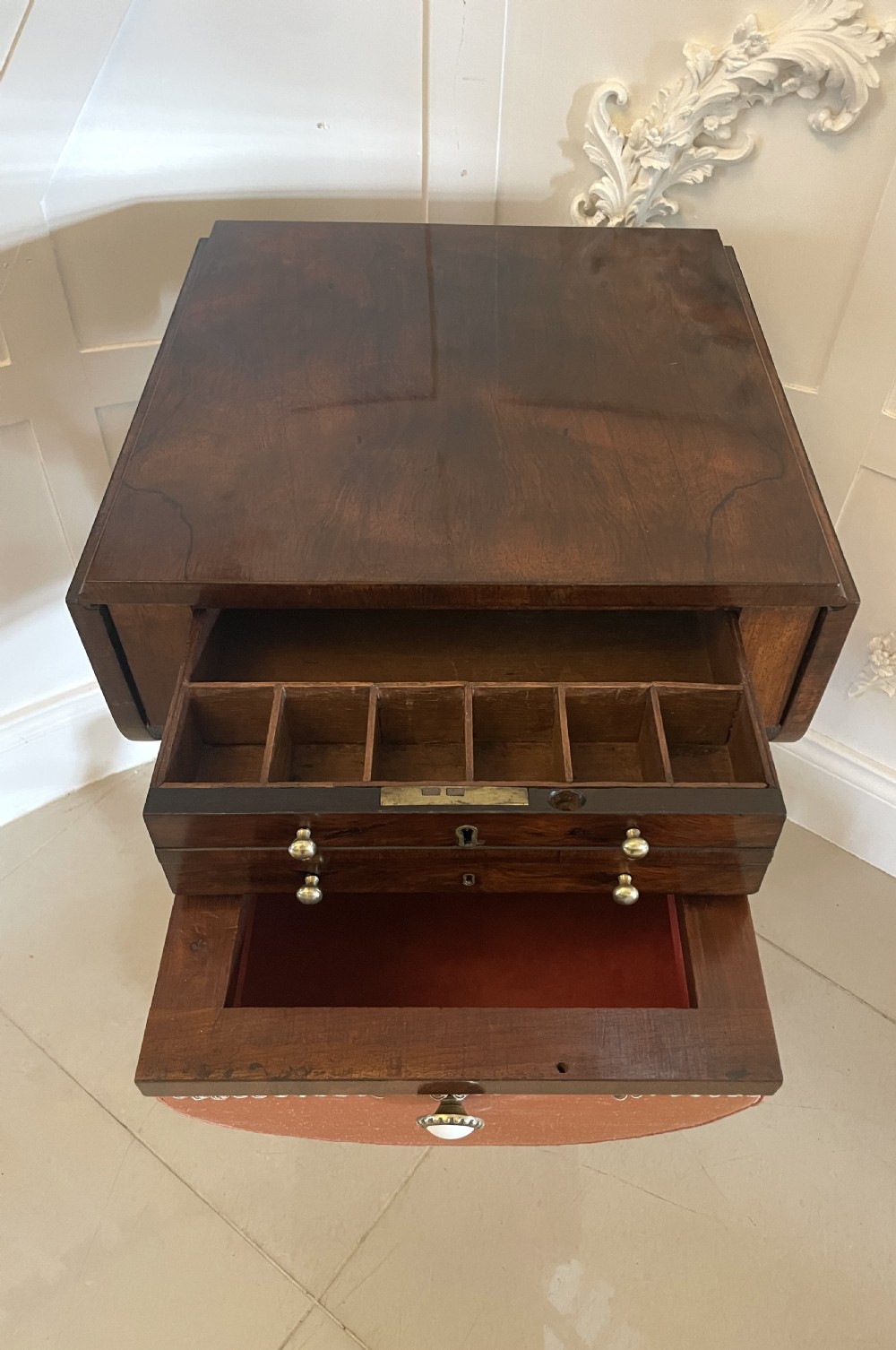 An antique Regency rosewood sewing table featuring small drop leaves, drawers with brass handles, a terracotta fabric work basket below, and a pedestal base with four splayed legs on castors. View XIX